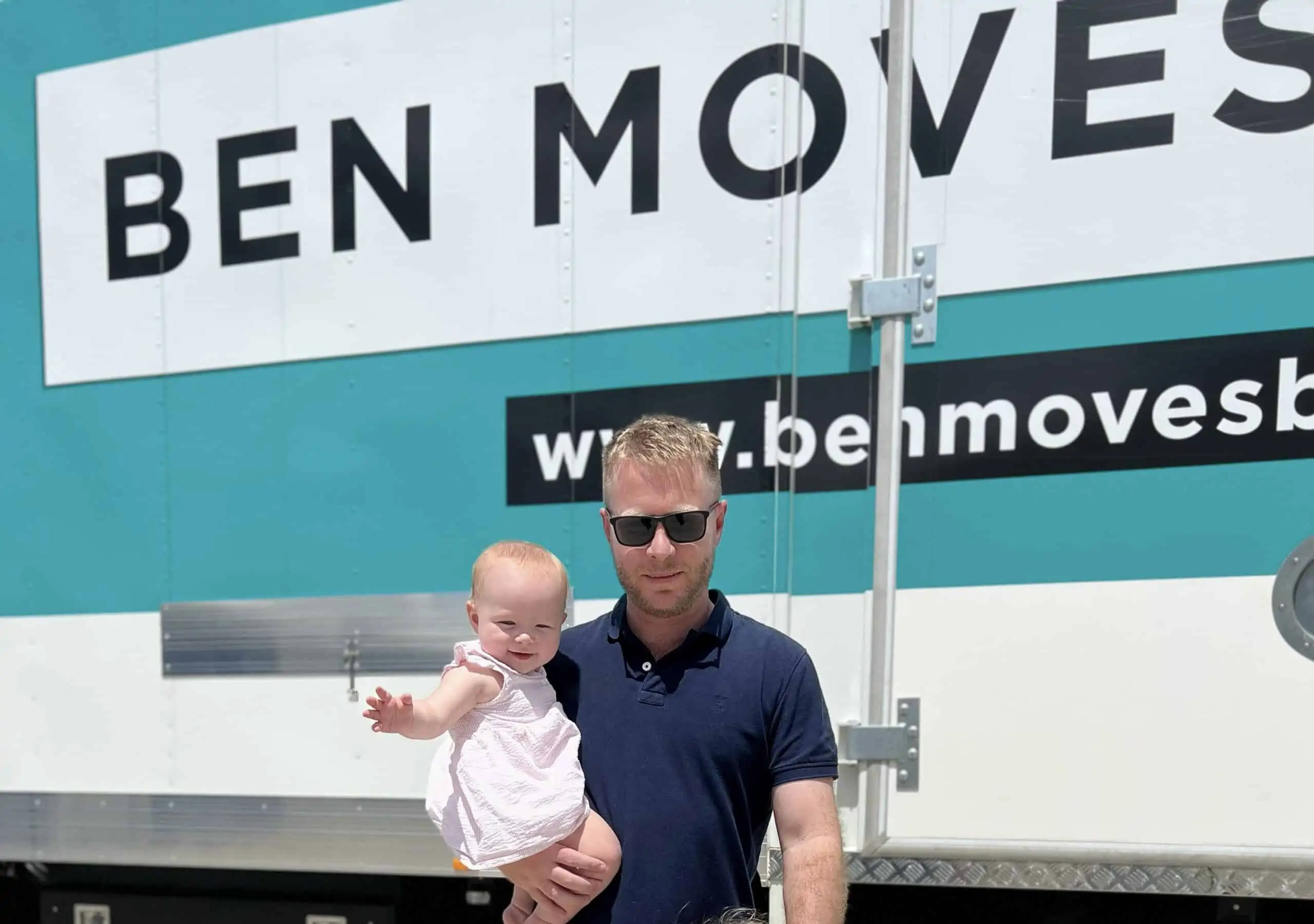 Ben Moves owner, Ben, holding his daughter in front of one of his moving trucks that is blue and white